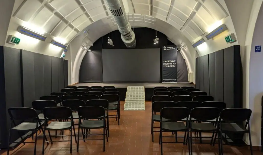 Sala koncertowa Żoliborskiego Domu Kultury - Fortu Sokolnickiego. Na środku sali znajduje się przejście prowadzące do sceny, po obu jego stronach ustawiono rzędy czarnych krzeseł dla publiczności. Scena ma czarne ściany i oświetlenie sceniczne. Pomieszczenie ma białe, łukowe sklepienie z widocznymi instalacjami wentylacyjnymi. Po bokach znajdują się oznaczenia wyjść ewakuacyjnych oraz symbol dostępności.