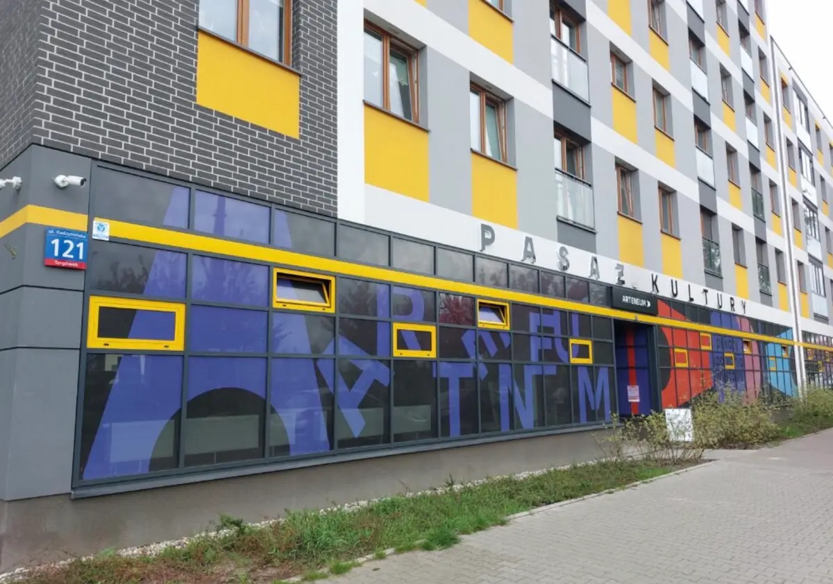Elewacja nowoczesnego budynku z dużym napisem „PASAŻ KULTURY” oraz „ARTENEUM” nad wejściem. Na rogu znajduje się niebieska tabliczka adresowa „ul. Radzymińska 121 Targówek”. Witryny na parterze pokryte są fioletowo-niebieską grafiką z żółtymi ramami okien, a elewacja wyższych pięter wykończona jest w barwach szarych, białych i żółtych. Przed budynkiem przebiega chodnik z kostki brukowej i wąski pas zieleni.